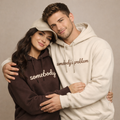 Couple hugging while wearing matching embroidered hoodies – dark brown hoodie with beige “somebody” embroidery and beige hoodie with brown “somebody’s problem” embroidery, premium couple hoodie set by Balaji Customi